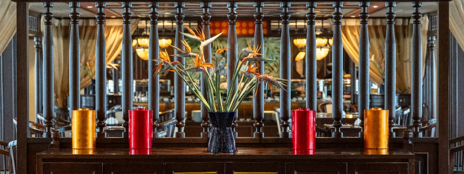 A dark wooden console holds a vase of bird-of-paradise flowers between red and orange cylindrical canisters, with turned wooden balusters and a softly lit restaurant or lounge with draped curtains behind.