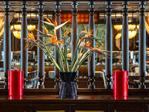 A dark wooden console holds a vase of bird-of-paradise flowers between red and orange cylindrical canisters, with turned wooden balusters and a softly lit restaurant or lounge with draped curtains behind.