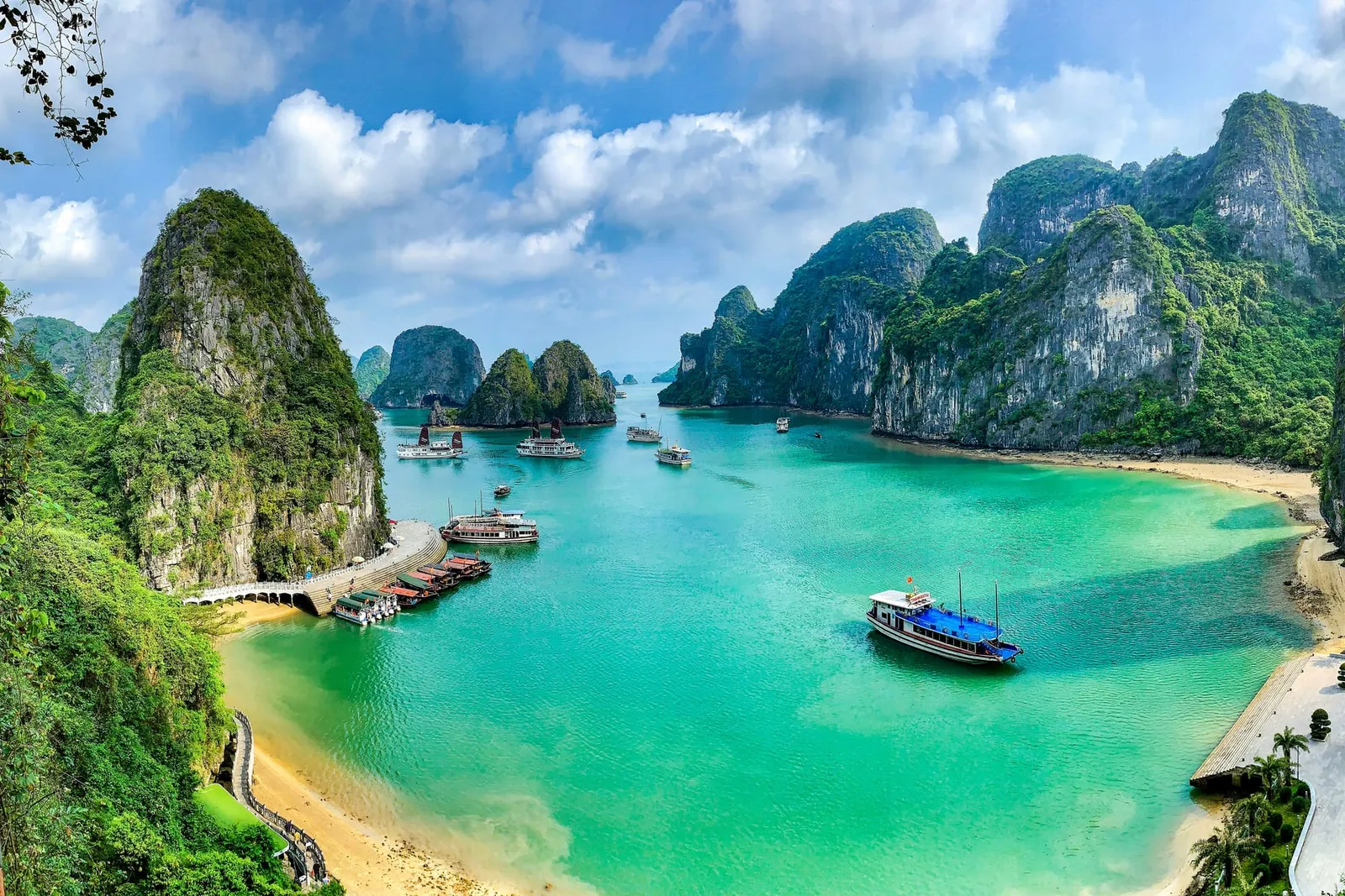 Panoramic view from an elevated cave viewpoint with a wooden railing, showing turquoise water, a sandy beach, a pier, several cruise boats, and tall limestone karsts covered in greenery under a blue sky with clouds.