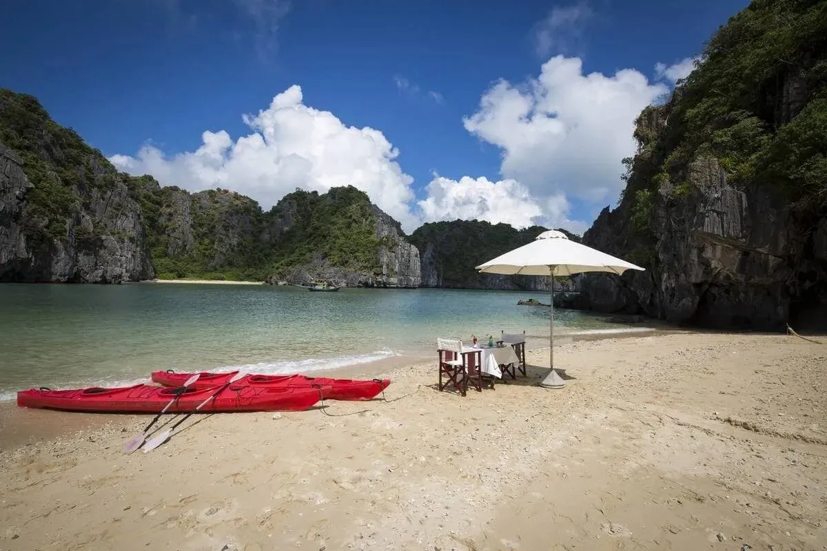 A secluded beach with red kayaks, dining setup under umbrella, surrounded by limestone karst cliffs and turquoise water.