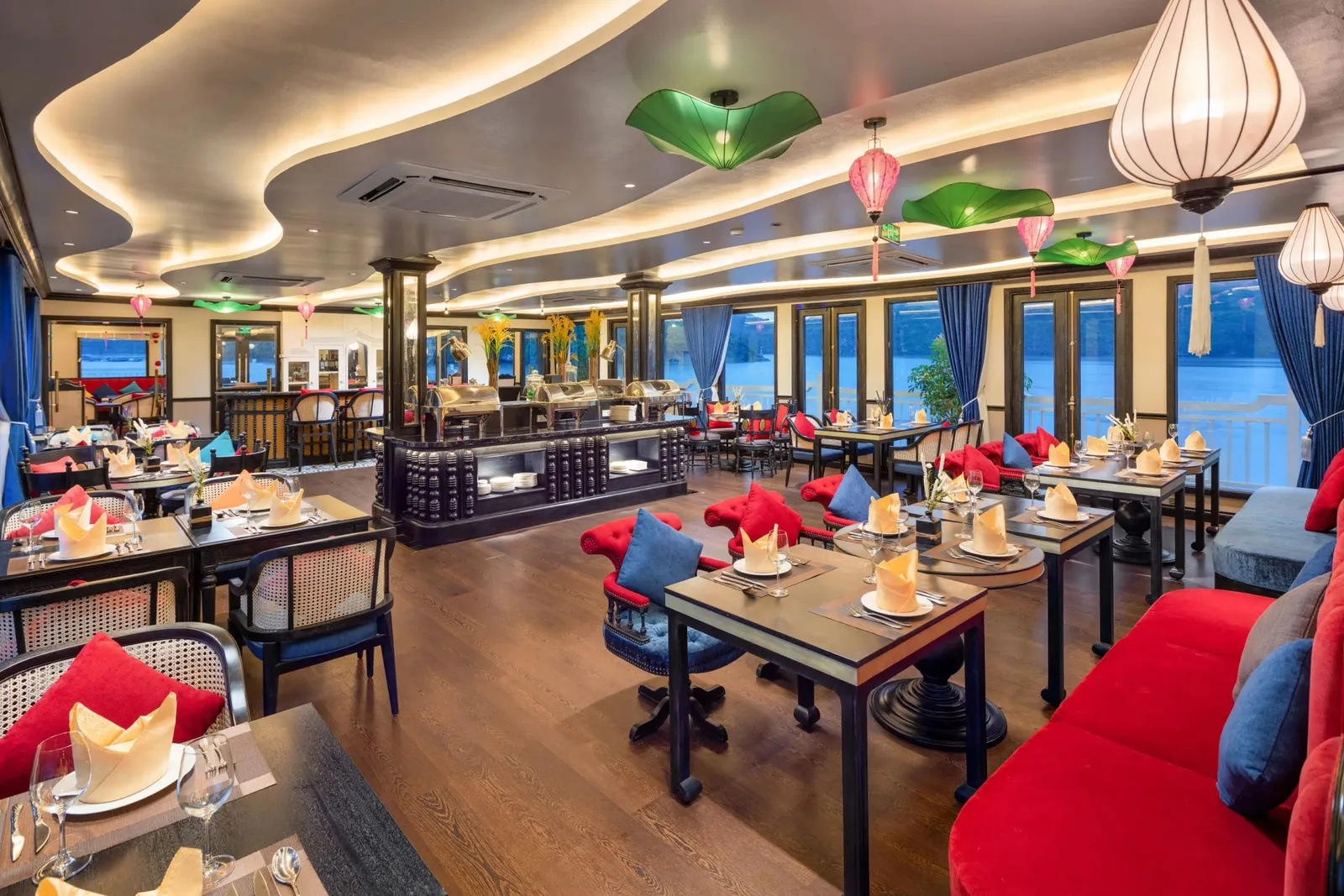 An interior restaurant dining room with set tables, red and blue seating, a buffet station, lantern-style lights and windows overlooking calm water.