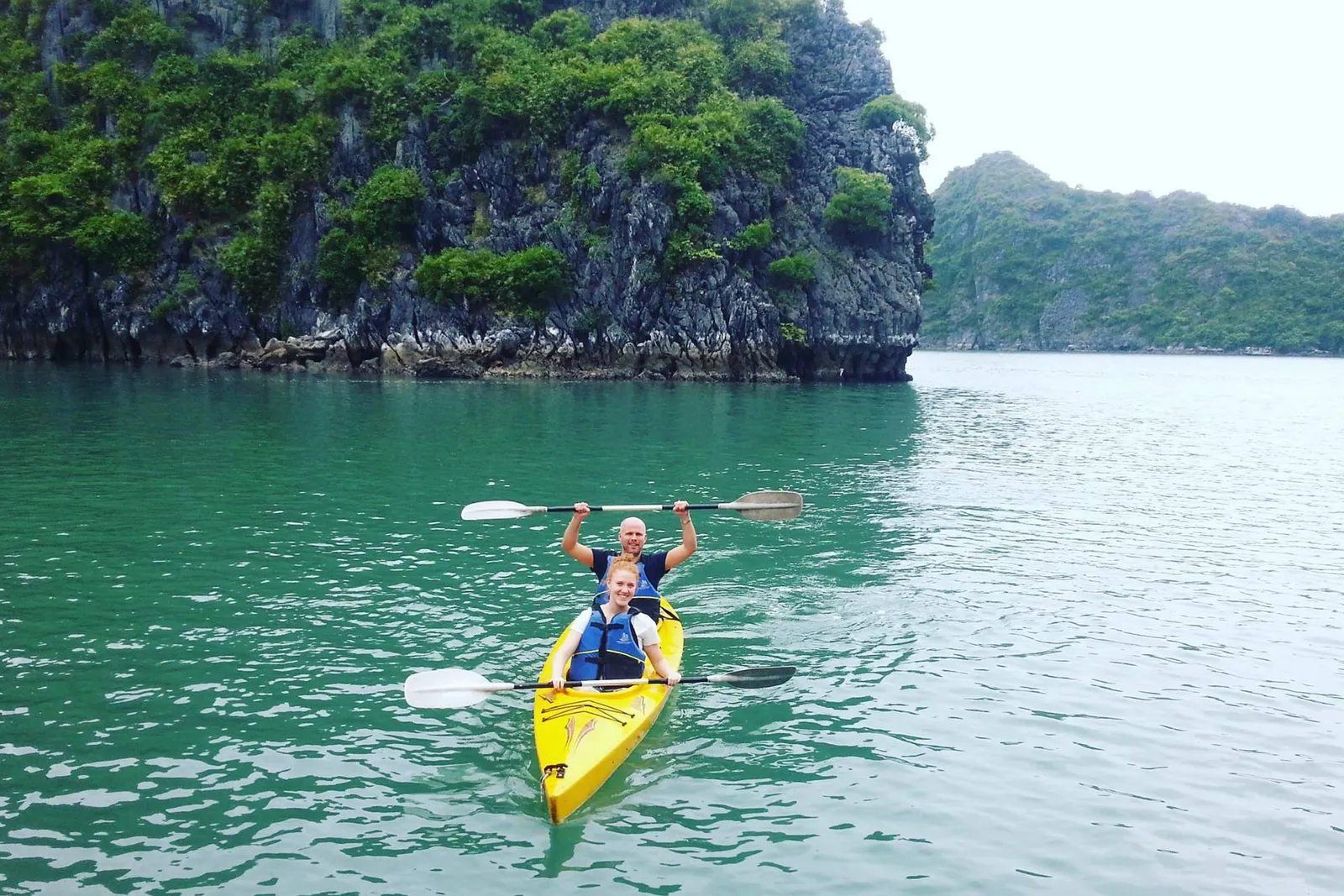 Two people in life vests paddle a yellow tandem kayak on green water, holding paddles up, with forested limestone karst cliffs behind them.