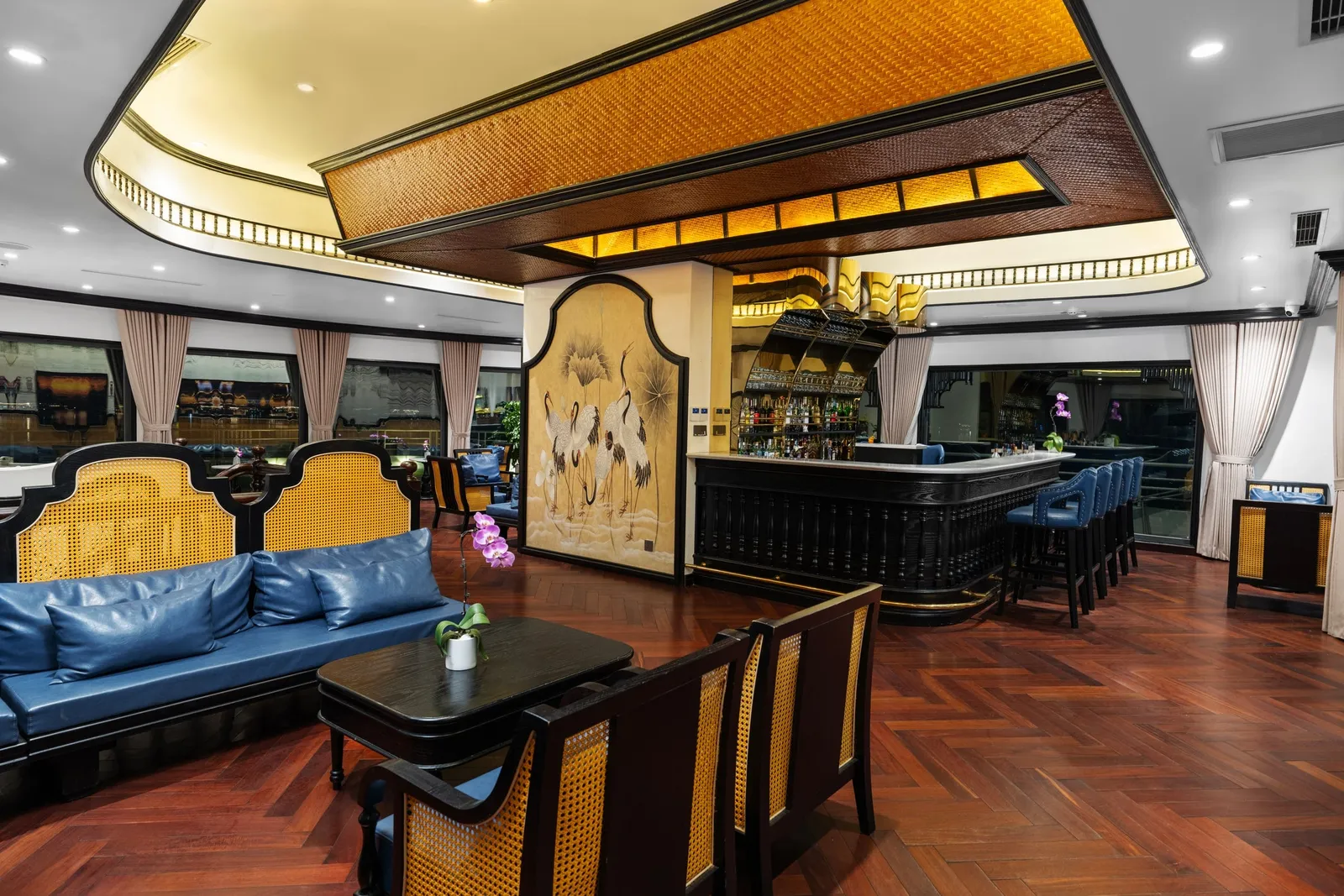An interior lounge bar with blue leather sofas, rattan-backed dark wood chairs, a curved black bar with blue stools, a crane mural panel, a woven ceiling feature and herringbone wood flooring.