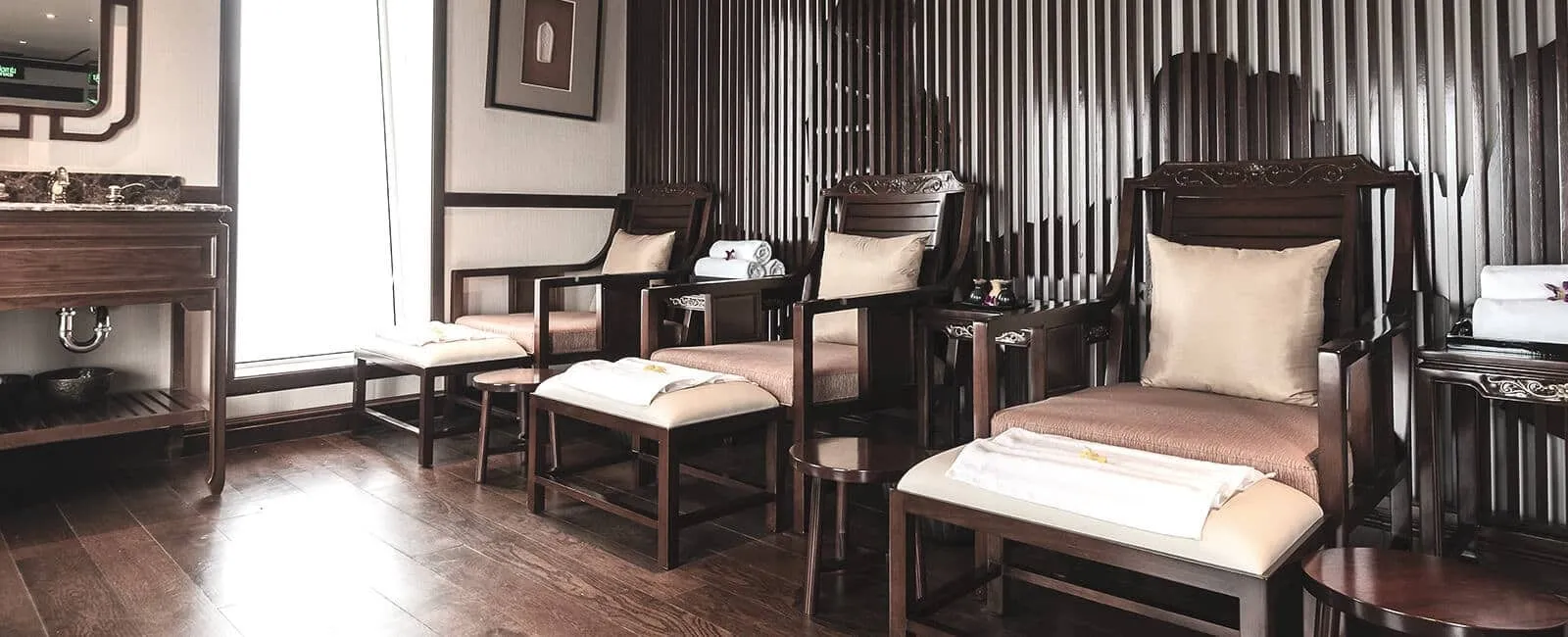 A spa or pedicure room with dark wood armchairs, footstools topped with folded white towels, a wooden vanity with a sink on the left, and a window letting in daylight.