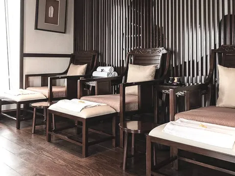 A spa or pedicure room with dark wood armchairs, footstools topped with folded white towels, a wooden vanity with a sink on the left, and a window letting in daylight.