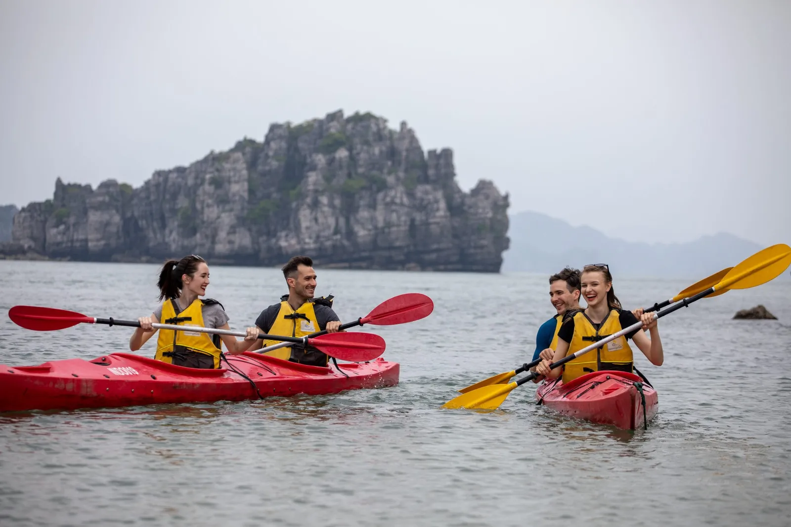 Four people in yellow life jackets kayaking in red and pink kayaks with dramatic limestone karst formations in the background.