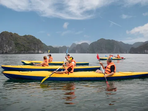 Several people wearing orange life jackets paddle yellow and blue kayaks on calm water with limestone karst formations in the background.