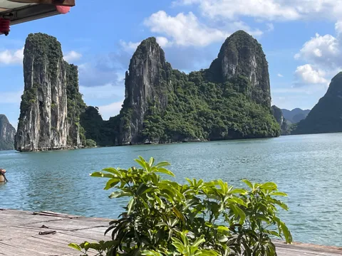 A wooden deck with a potted plant in the foreground overlooks calm water and tall limestone karst formations under a partly cloudy sky.