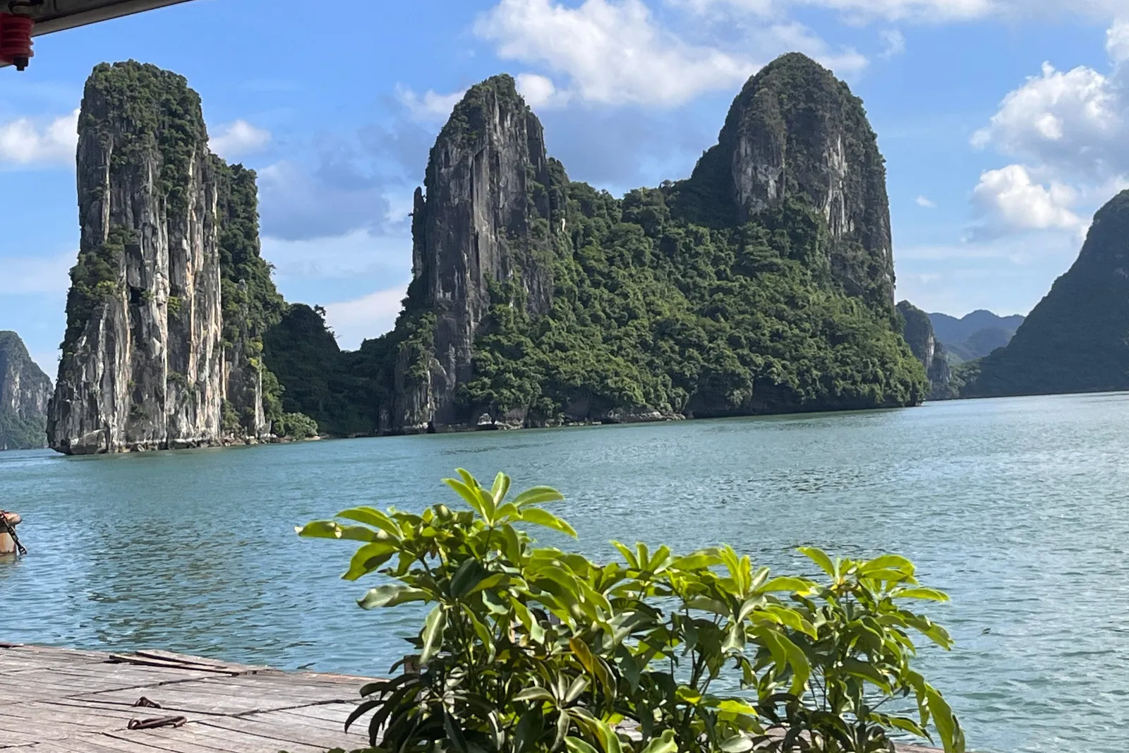 A wooden deck with a potted plant in the foreground overlooks calm water and tall limestone karst formations under a partly cloudy sky.