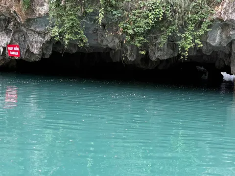 Turquoise water in front of a limestone cliff with a cave entrance and a red sign with white text on the rock face.