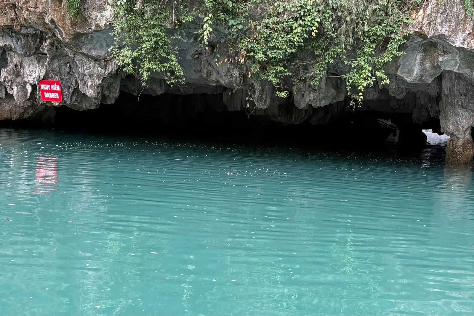 Turquoise water in front of a limestone cliff with a cave entrance and a red sign with white text on the rock face.