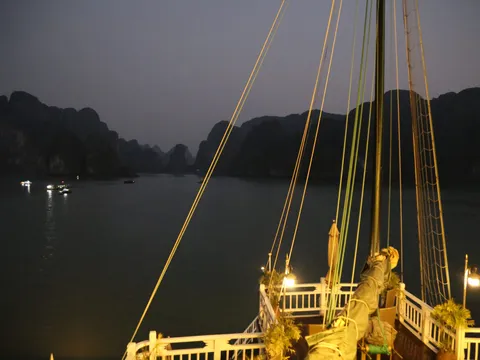 A twilight view from a traditional junk boat's deck showing illuminated rigging and limestone karsts across calm water.