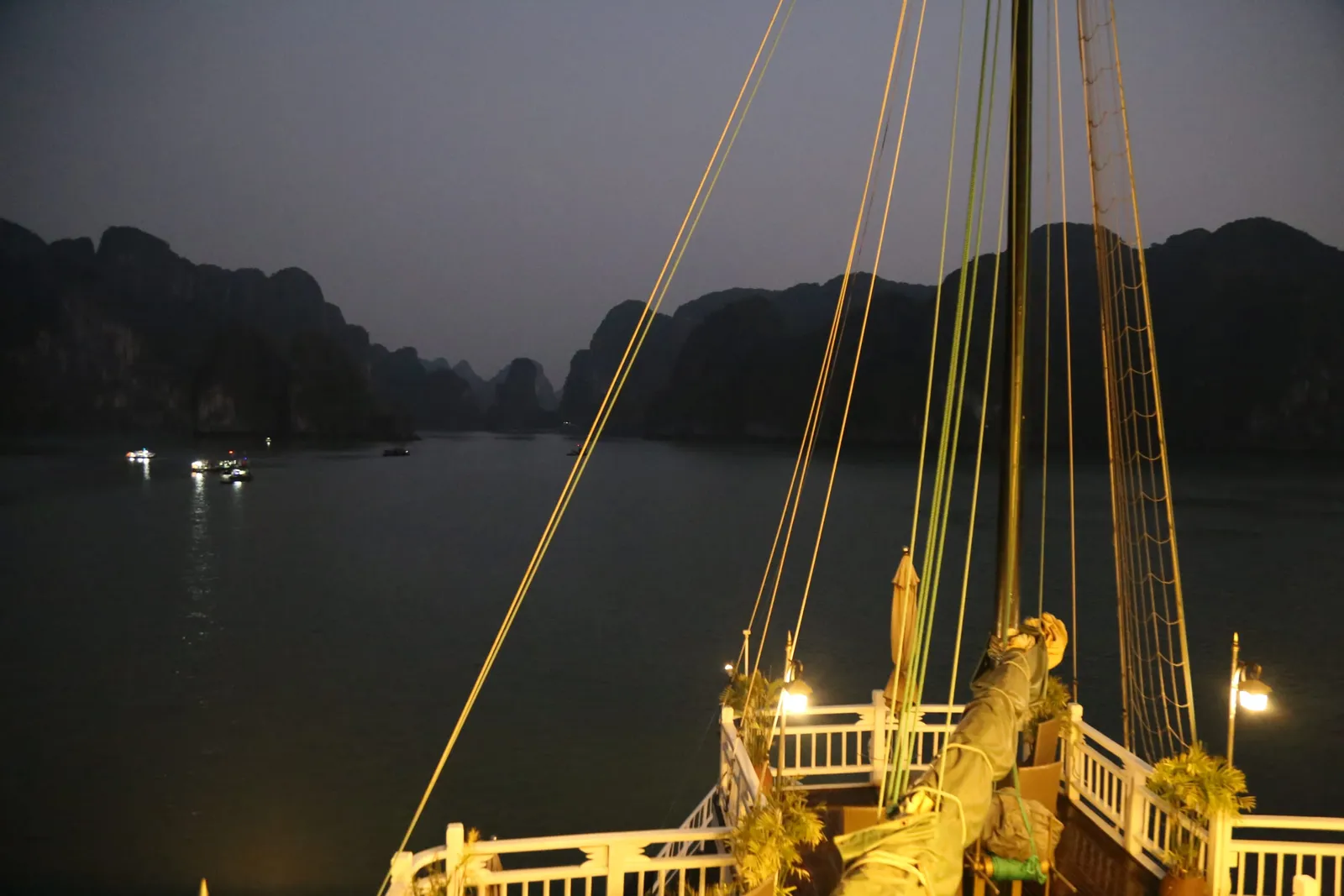 A twilight view from a traditional junk boat's deck showing illuminated rigging and limestone karsts across calm water.