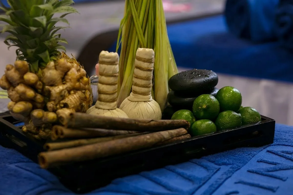 A black tray of spa items (herbal compress balls, ginger, pineapple top, lemongrass, cinnamon sticks, black stones, limes) on a blue towel with embossed lettering.