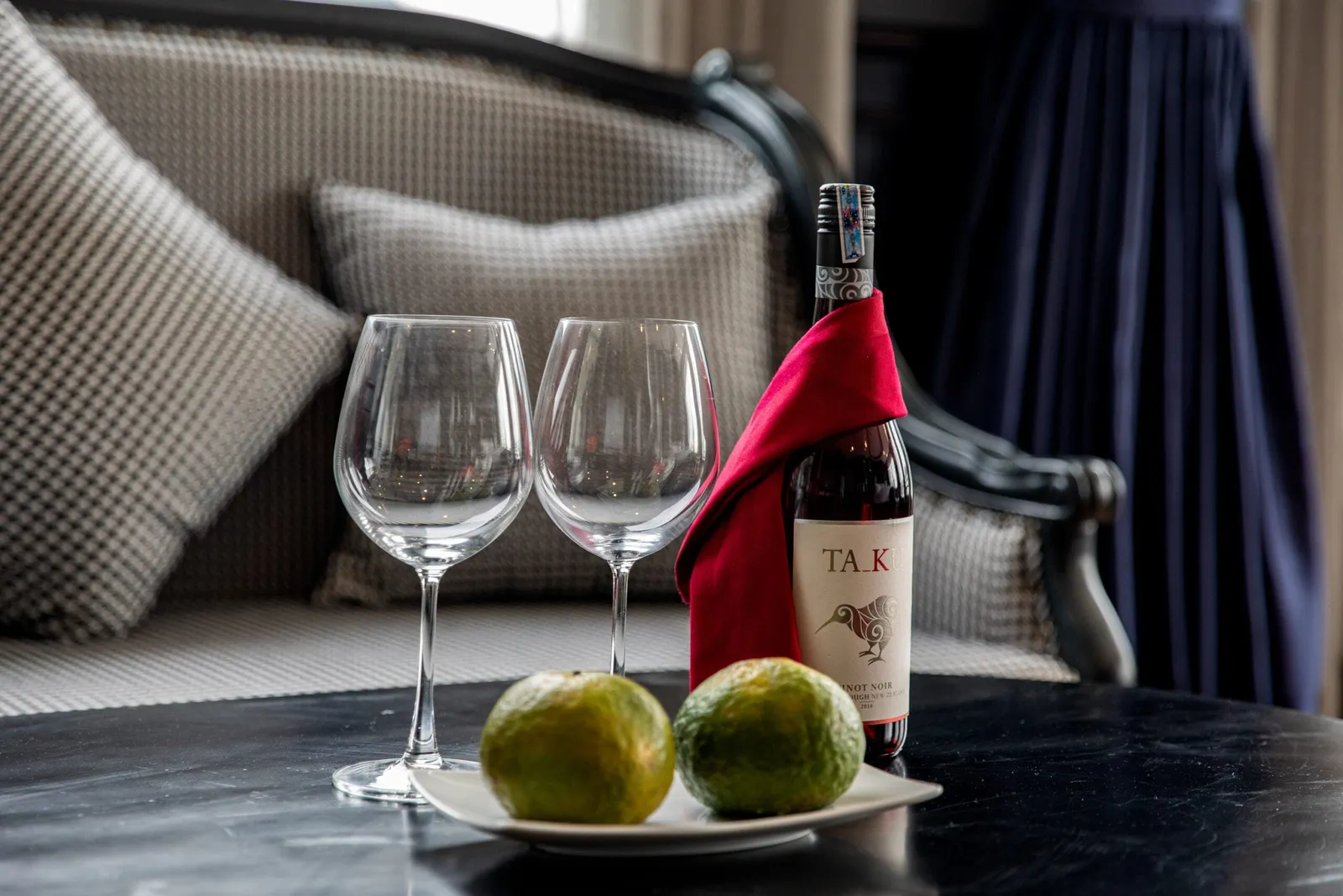 Two empty wine glasses, a Pinot Noir bottle wrapped in a red cloth and a plate of two green citrus fruits on a dark table in front of a houndstooth-cushioned sofa and navy curtains.
