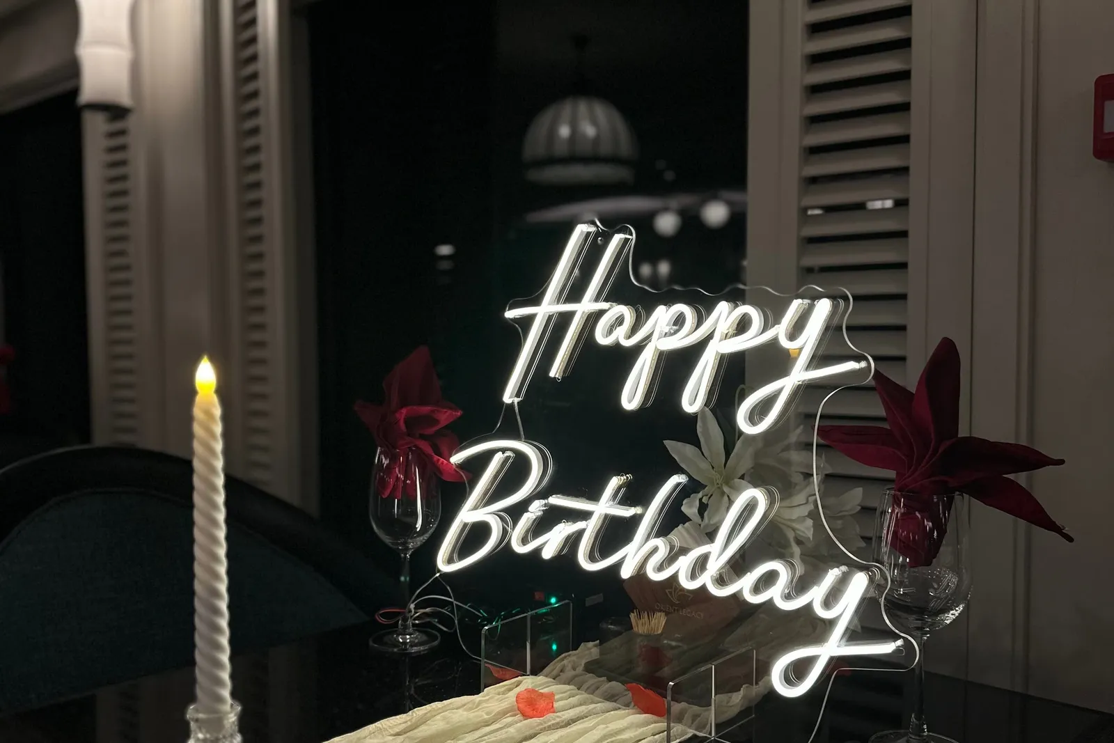 A lit neon 'Happy Birthday' sign stands on a dark table set with a cream runner, a twisted candle, wine glasses with red napkins and red petals, in an indoor room with louvered shutters at night.