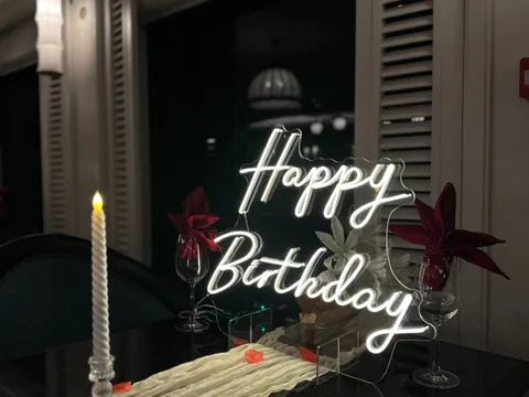 A lit neon 'Happy Birthday' sign stands on a dark table set with a cream runner, a twisted candle, wine glasses with red napkins and red petals, in an indoor room with louvered shutters at night.