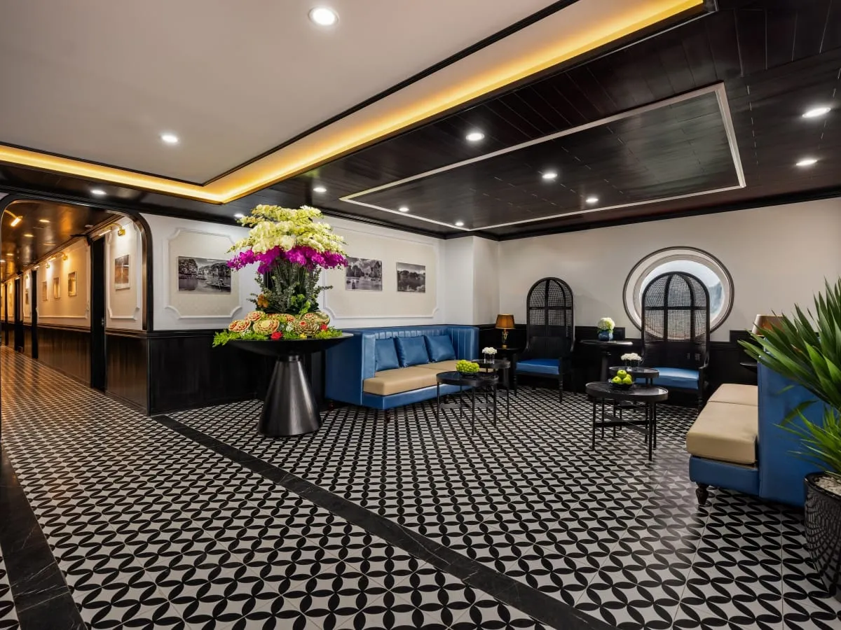 A lounge interior with black-and-white patterned tile floor, blue banquette seating, a large orchid and carved-fruit display, framed black-and-white bay photos on the wall, a round porthole-style window and a corridor to the left.