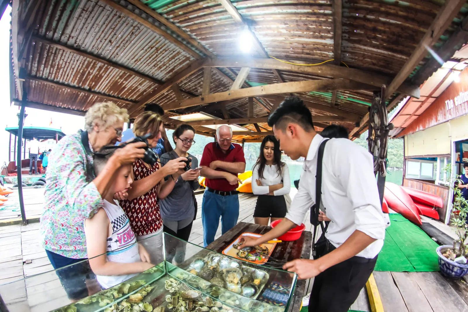 A young man in a white shirt shows shellfish from a glass tank to a group of tourists photographing him on a floating platform under a corrugated roof, with kayaks and green water behind.