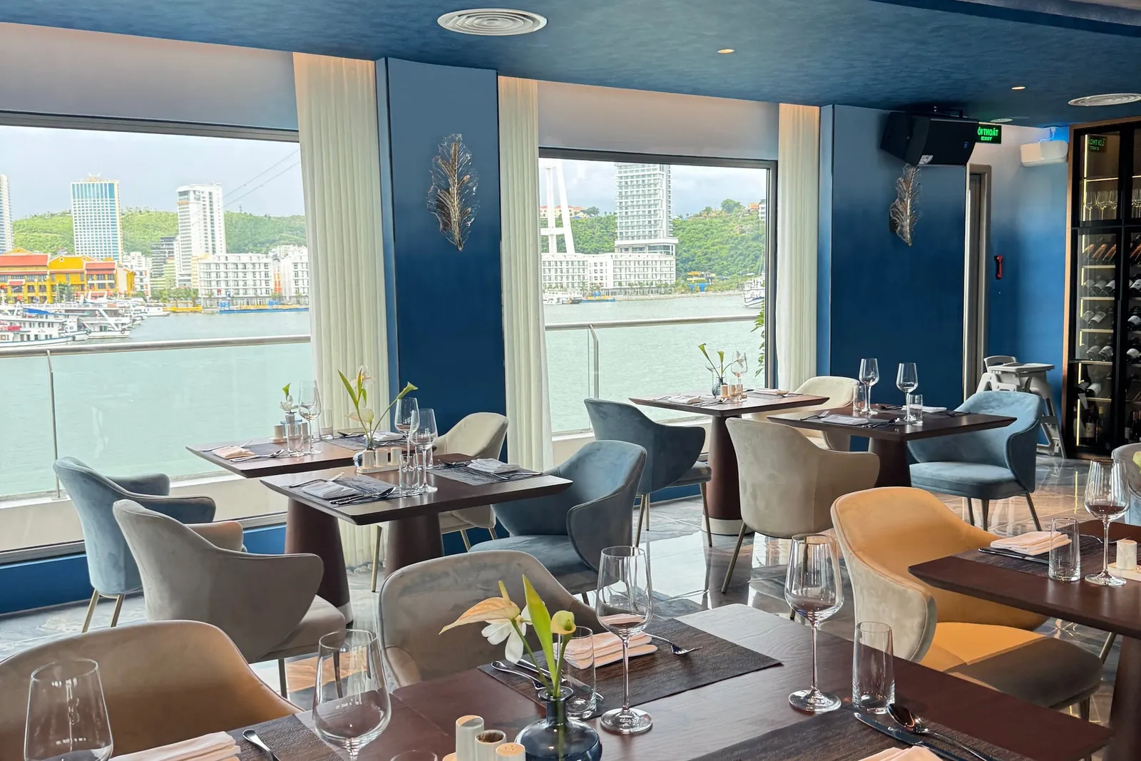 A blue-toned dining room with set tables, velvet chairs and a wine cabinet, with large windows looking out to the water, a waterfront of high-rise buildings, green hills and moored cruise boats.