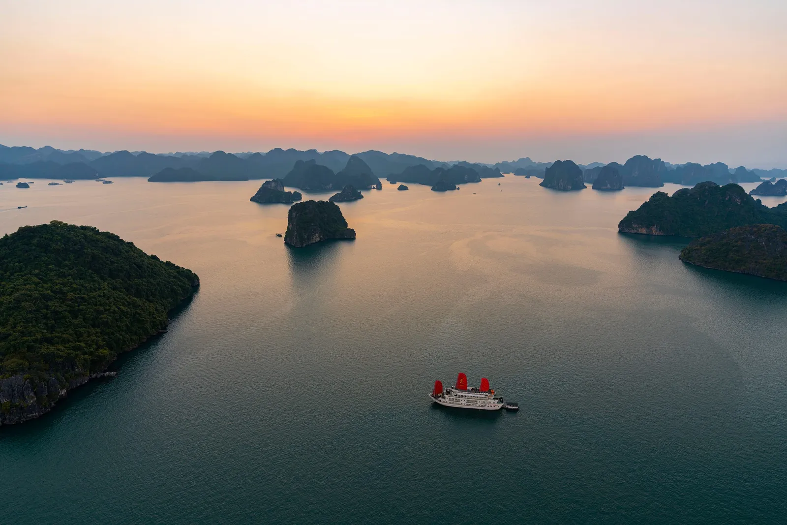 An aerial view at sunset of a white cruise boat with red sails on calm water, surrounded by forested limestone karst islands under an orange and pink sky.