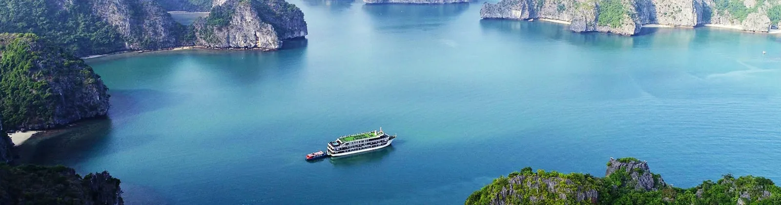 Elevated view of a multi-deck cruise boat with a small red tender anchored in turquoise water surrounded by forested limestone karst islands.