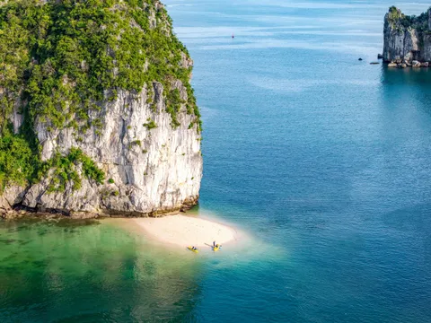 Aerial view of a small beach with two kayakers between tall limestone karsts and turquoise water.