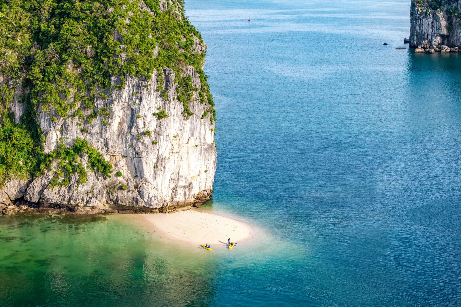 Aerial view of a small beach with two kayakers between tall limestone karsts and turquoise water.
