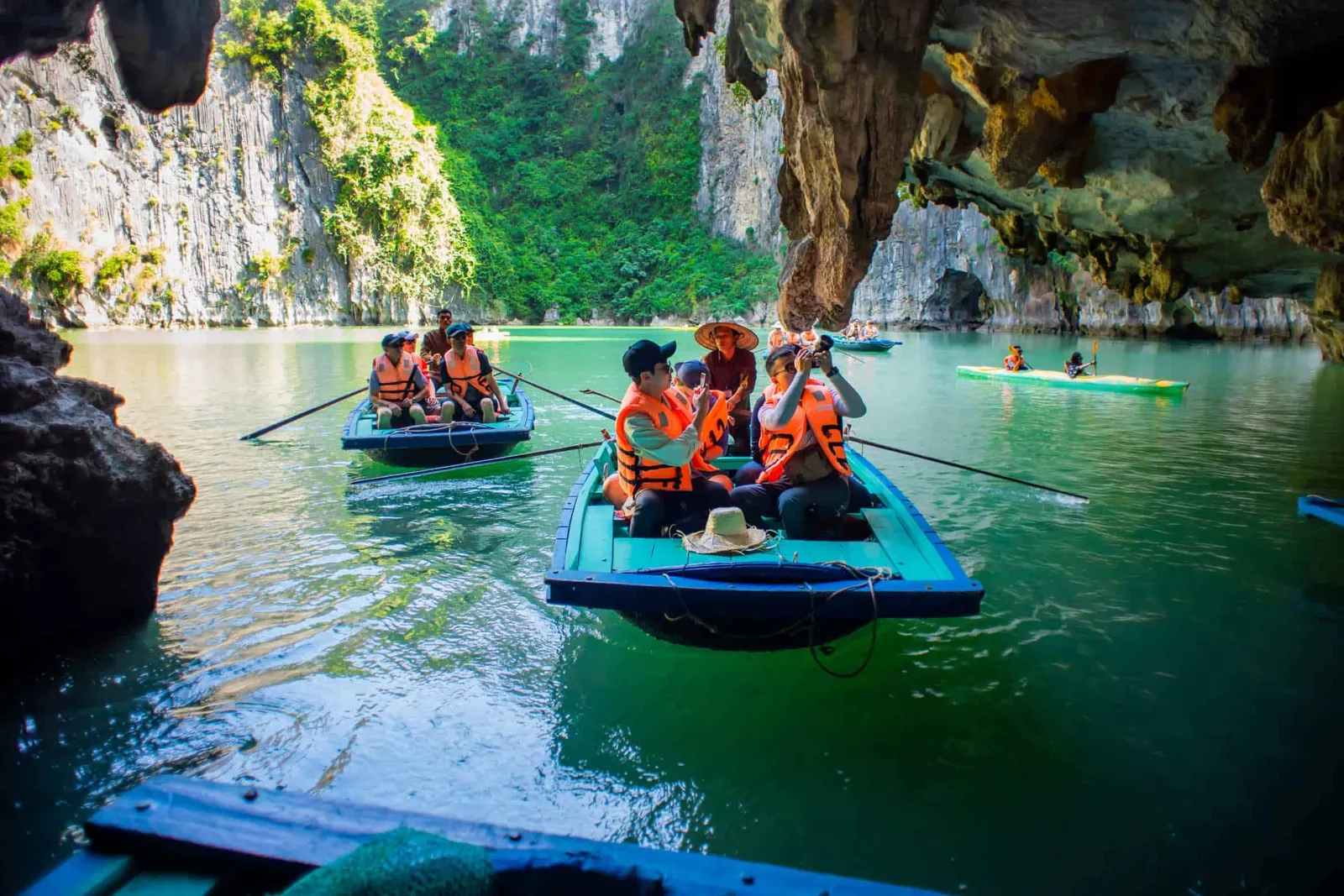 Tourists in orange life jackets ride small rowboats with a rower in a conical hat through emerald water beneath a limestone cave overhang, with green-covered karst cliffs and a kayak in the background, photographed from another small boat whose blue edge is in the foreground.