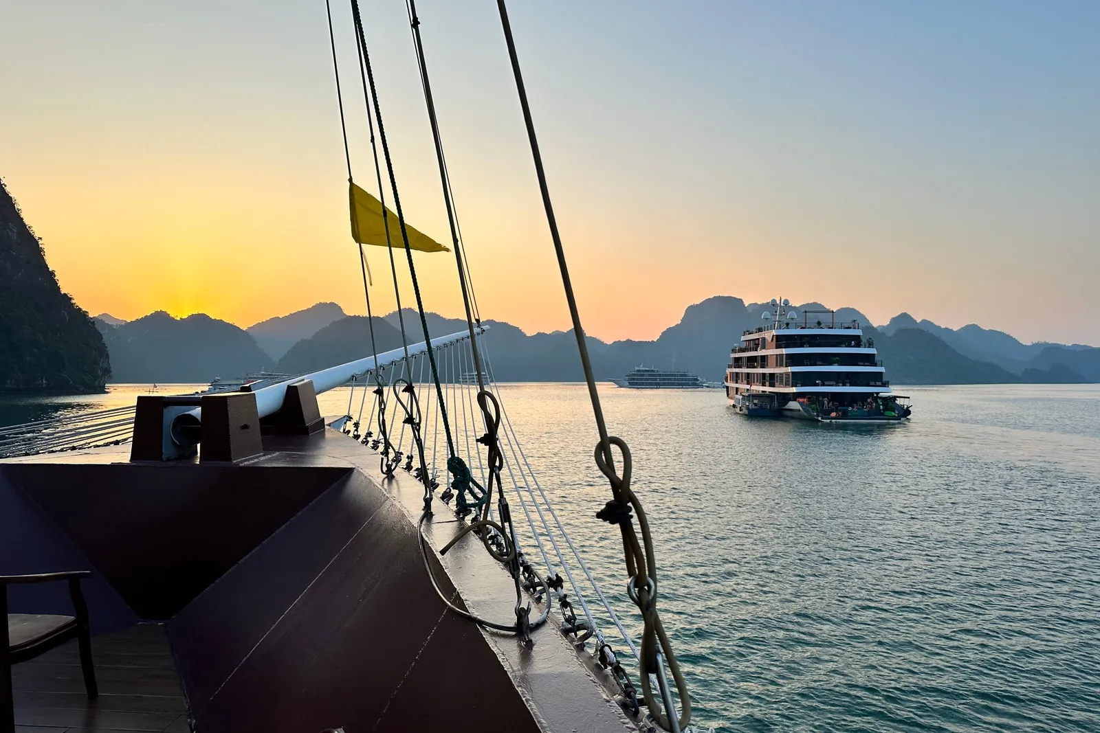 A view from the deck of a boat showing another cruise ship in calm waters surrounded by karst mountains at sunset.