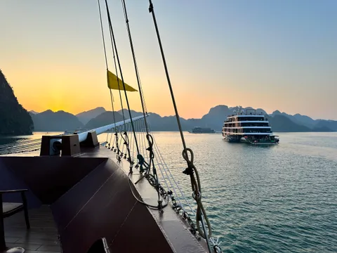 A view from the deck of a boat showing another cruise ship in calm waters surrounded by karst mountains at sunset.