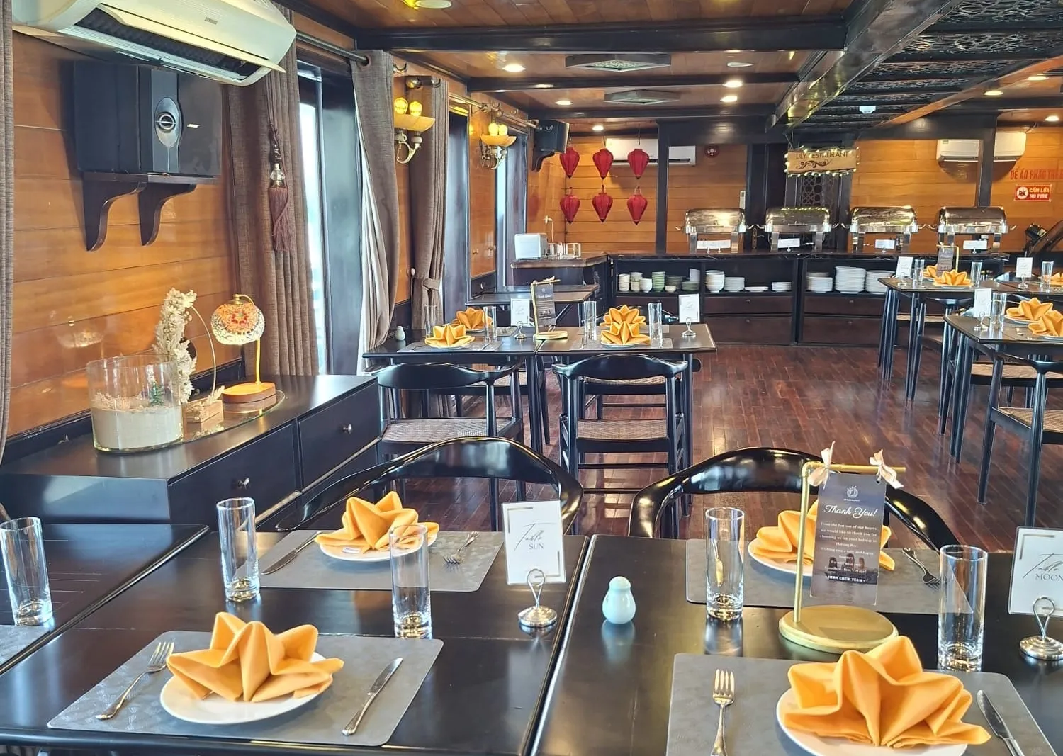 A wood-paneled dining room with black tables set with orange folded napkins, table number cards reading Sun and Moon, a buffet line with chafing dishes at the back, red lanterns, and a wall-mounted air conditioner.