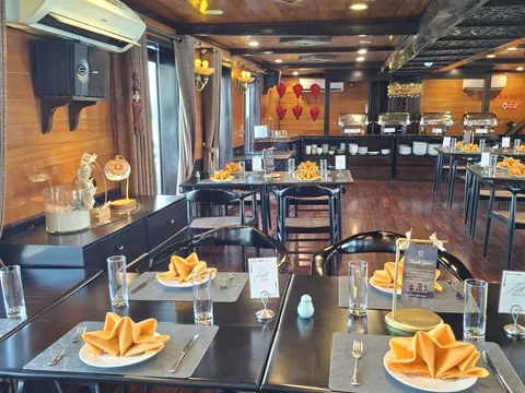 A wood-paneled dining room with black tables set with orange folded napkins, table number cards reading Sun and Moon, a buffet line with chafing dishes at the back, red lanterns, and a wall-mounted air conditioner.
