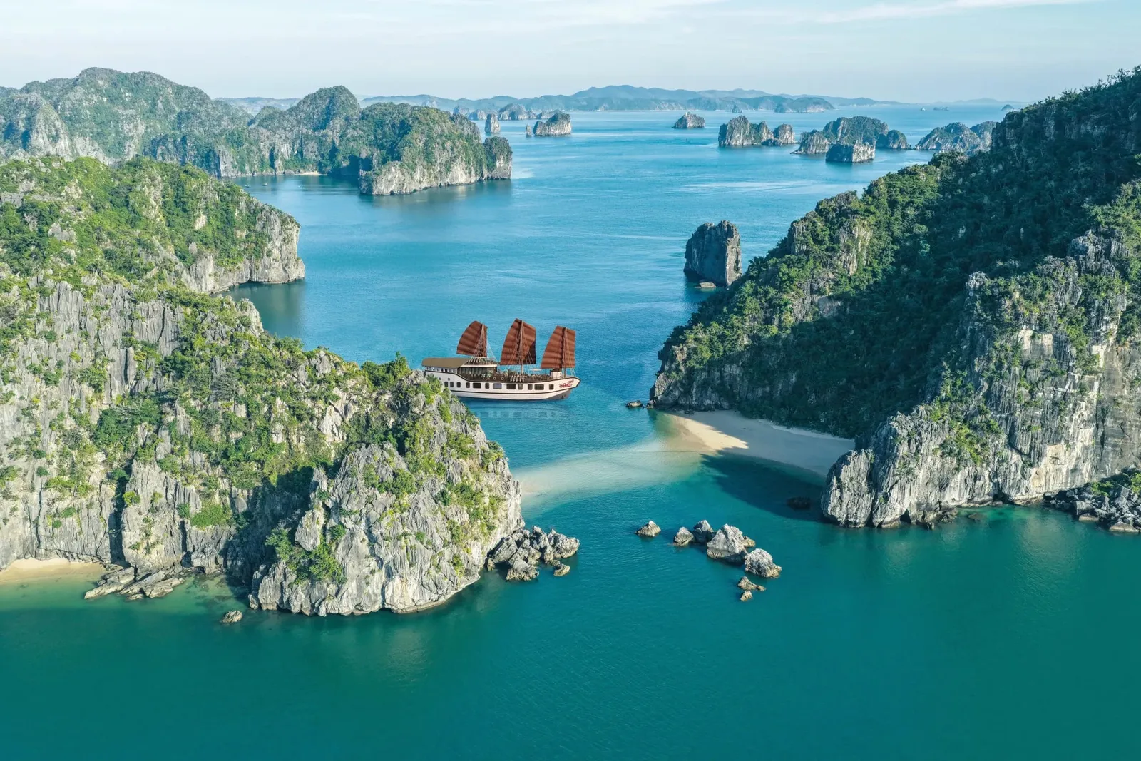 Aerial view of a white junk-style boat with three red-brown sails in turquoise water between green limestone karst islands, with small sandy beaches in the foreground.