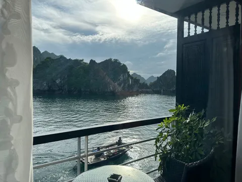 View from a boat cabin or deck showing limestone karsts rising from calm water with curtains, railings, and a small rowing boat visible in the foreground.