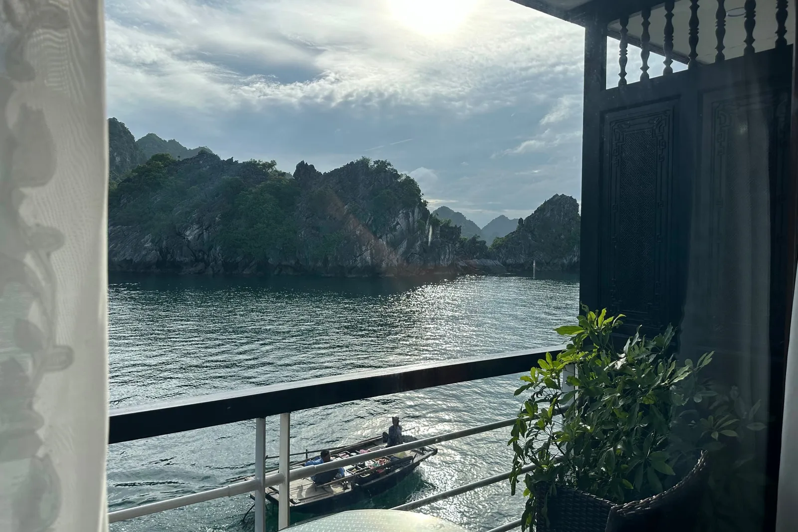 View from a boat cabin or deck showing limestone karsts rising from calm water with curtains, railings, and a small rowing boat visible in the foreground.