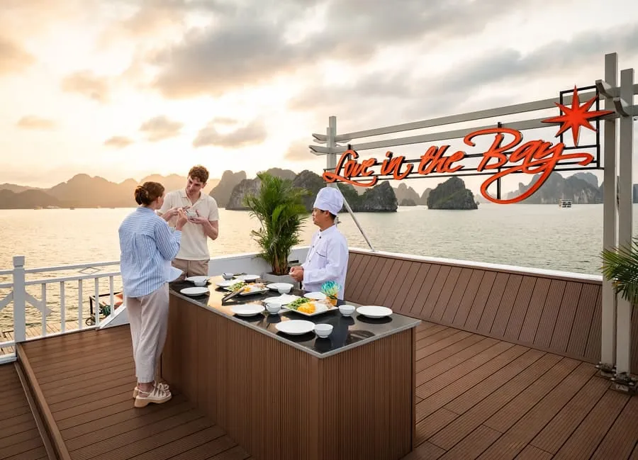 A chef serving two guests at an open-air food counter on deck at sunset.
