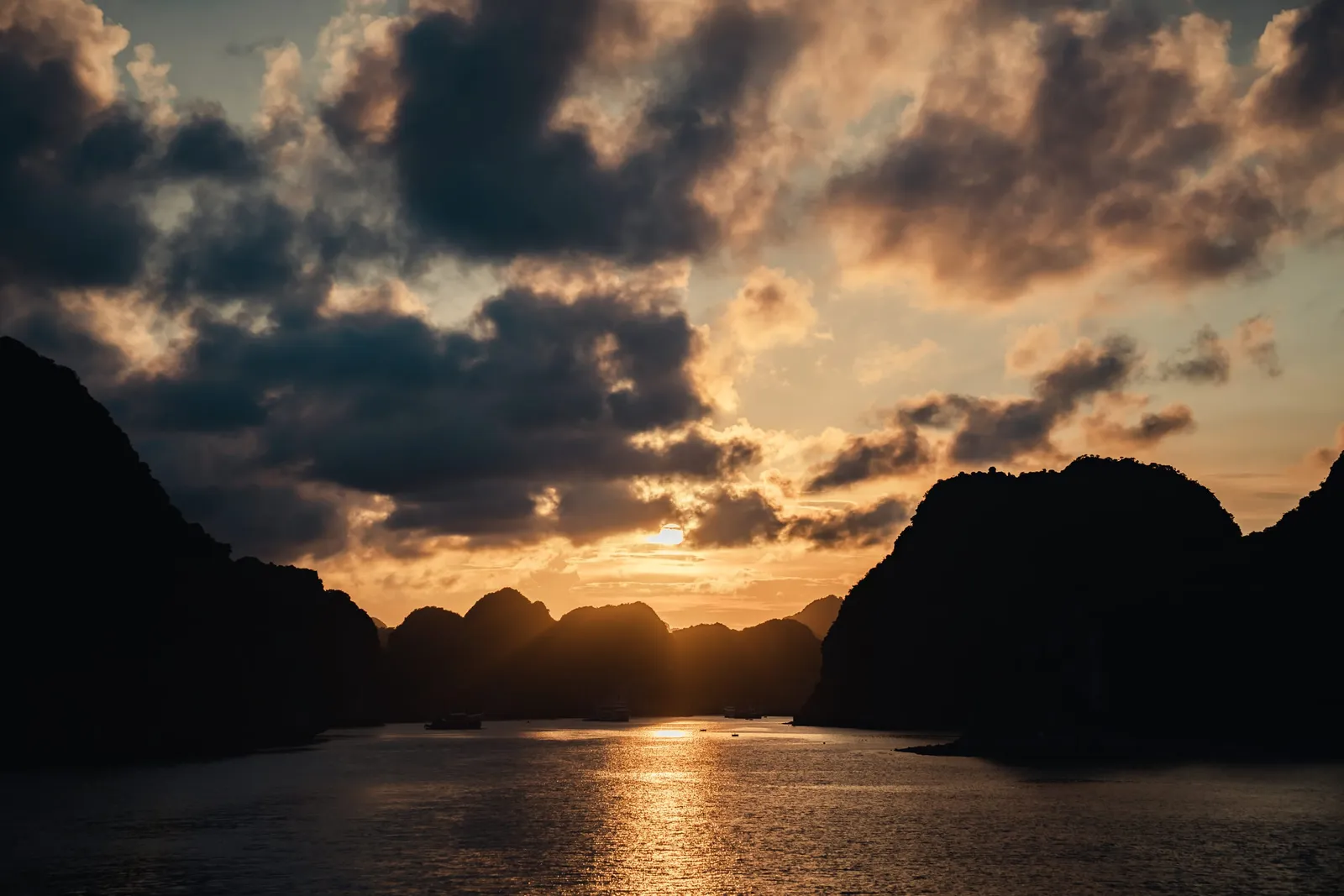 Sunset view through limestone karst islands with calm water reflecting golden light and silhouetted cruise boats in the distance.