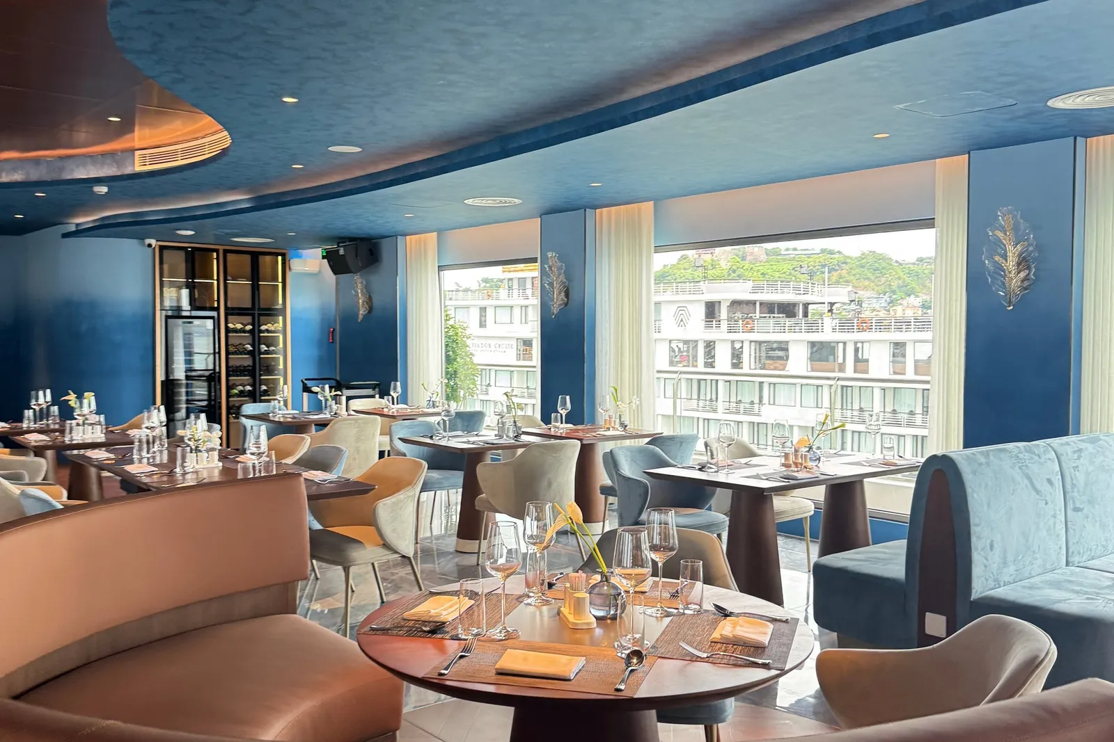 A curved blue-ceilinged restaurant interior with set tables and tan and blue seating, and a window showing a docked white cruise ship with a green hill behind it; a logo is visible on the neighbouring ship's hull.
