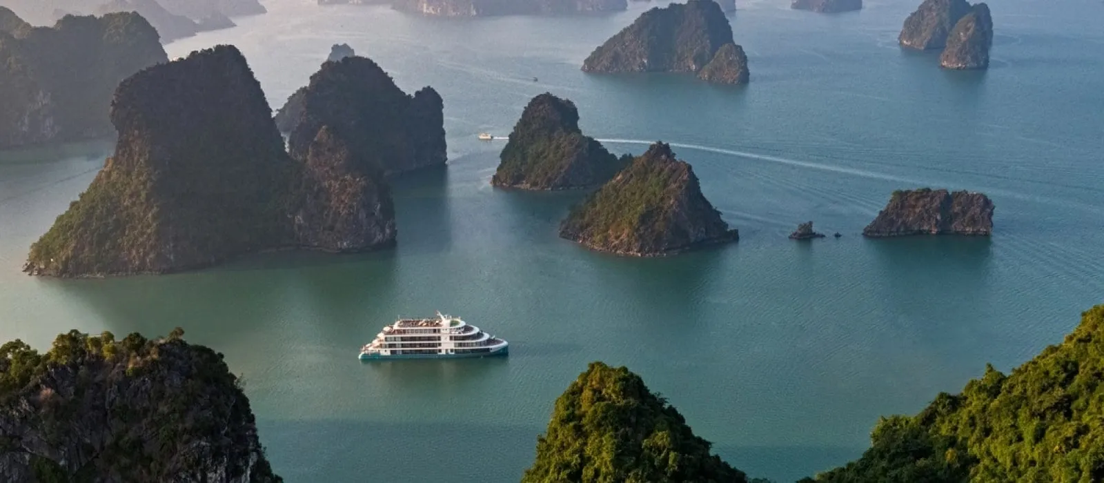 Aerial view of limestone karst islands in green water with a small white multi-deck cruise ship in the center and a distant boat leaving a wake.