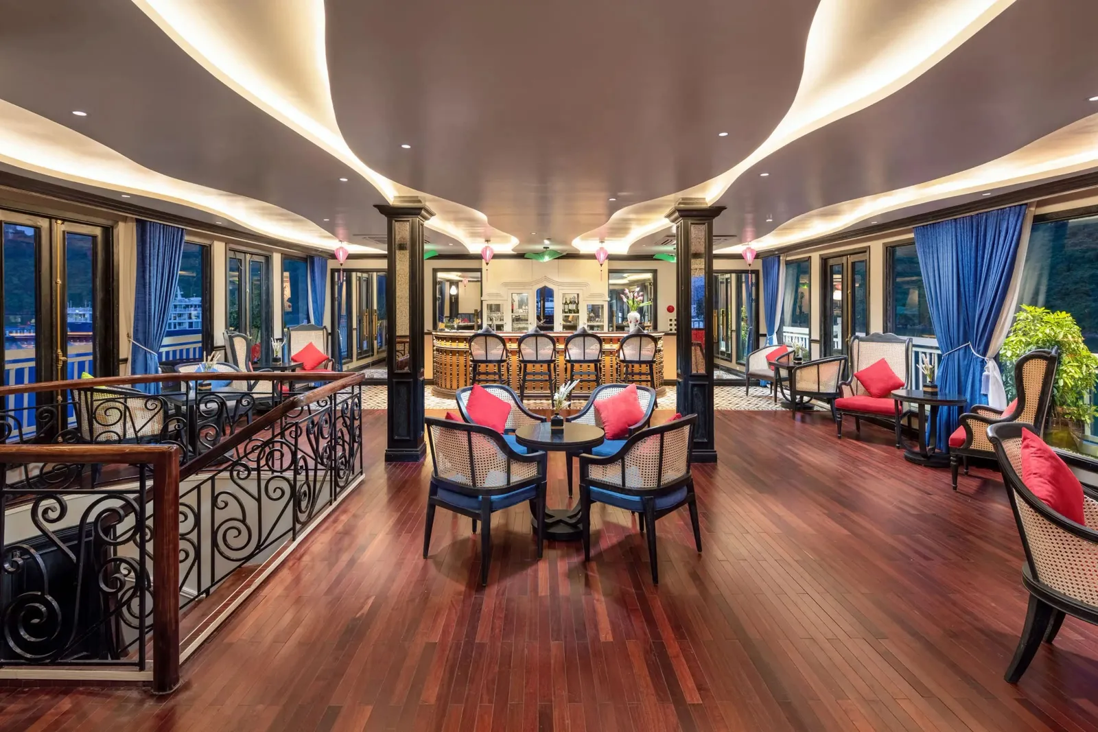 An interior lounge with a bar, rattan chairs with red and blue cushions, blue curtains, a dark hardwood floor, a wrought-iron stair railing and a glowing curved ceiling, with limestone cliffs and another vessel visible through the windows.