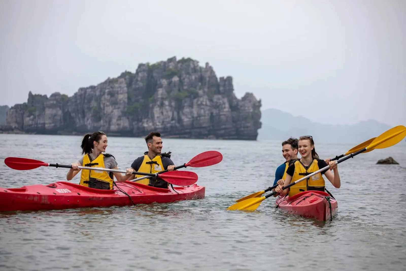 Four guests in life jackets paddling red kayaks in front of a limestone island.