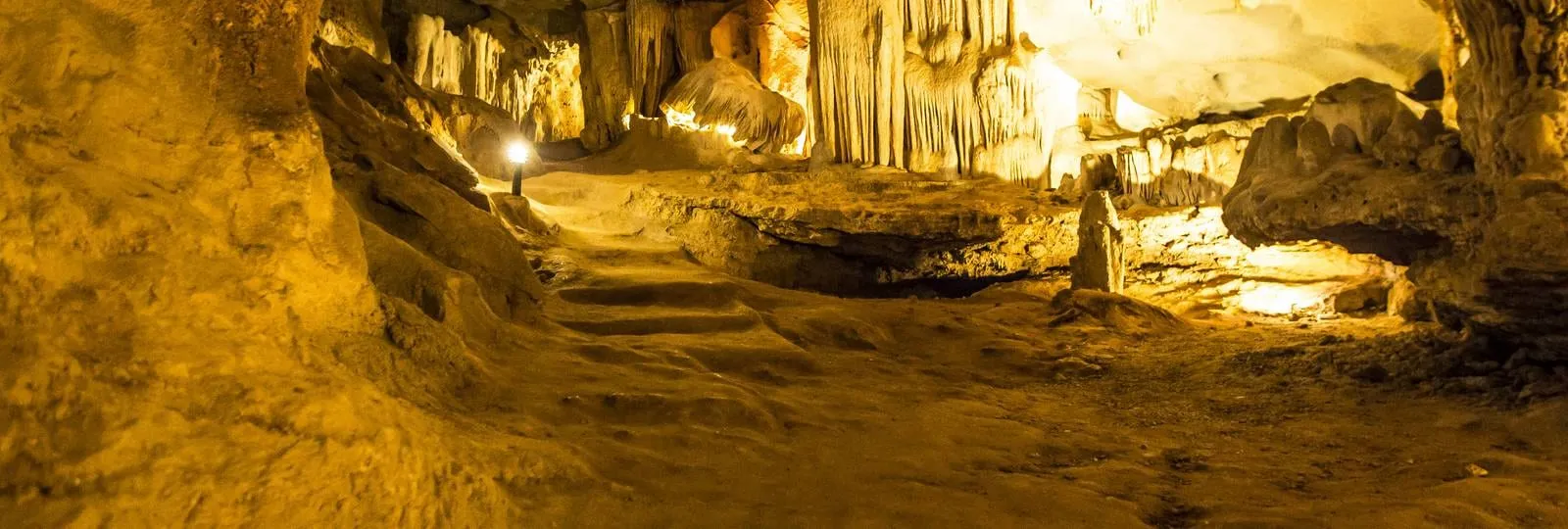 Interior of a limestone cave with stalactites and stalagmites, lit by warm artificial lighting along a walkway.