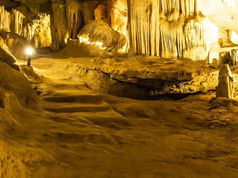 Interior of a limestone cave with stalactites and stalagmites, lit by warm artificial lighting along a walkway.