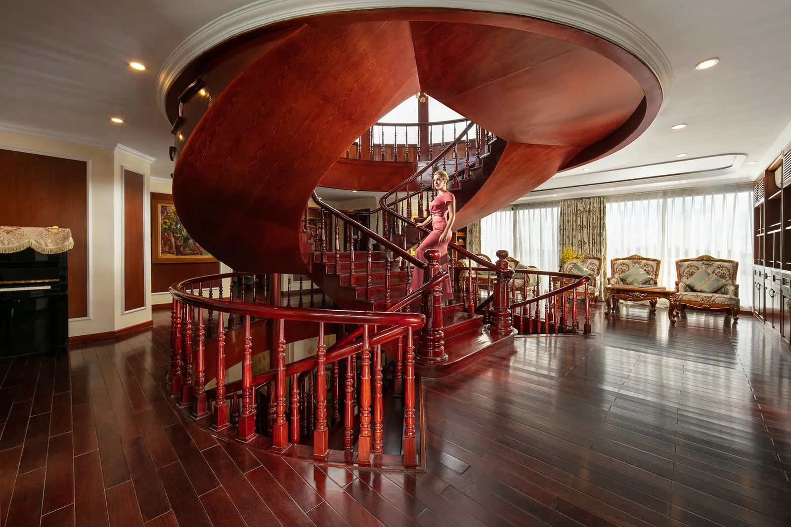 A woman in a pink dress stands on a red-wood spiral staircase in a large lounge with dark wood floors, ornate armchairs, sheer curtains and a black piano.