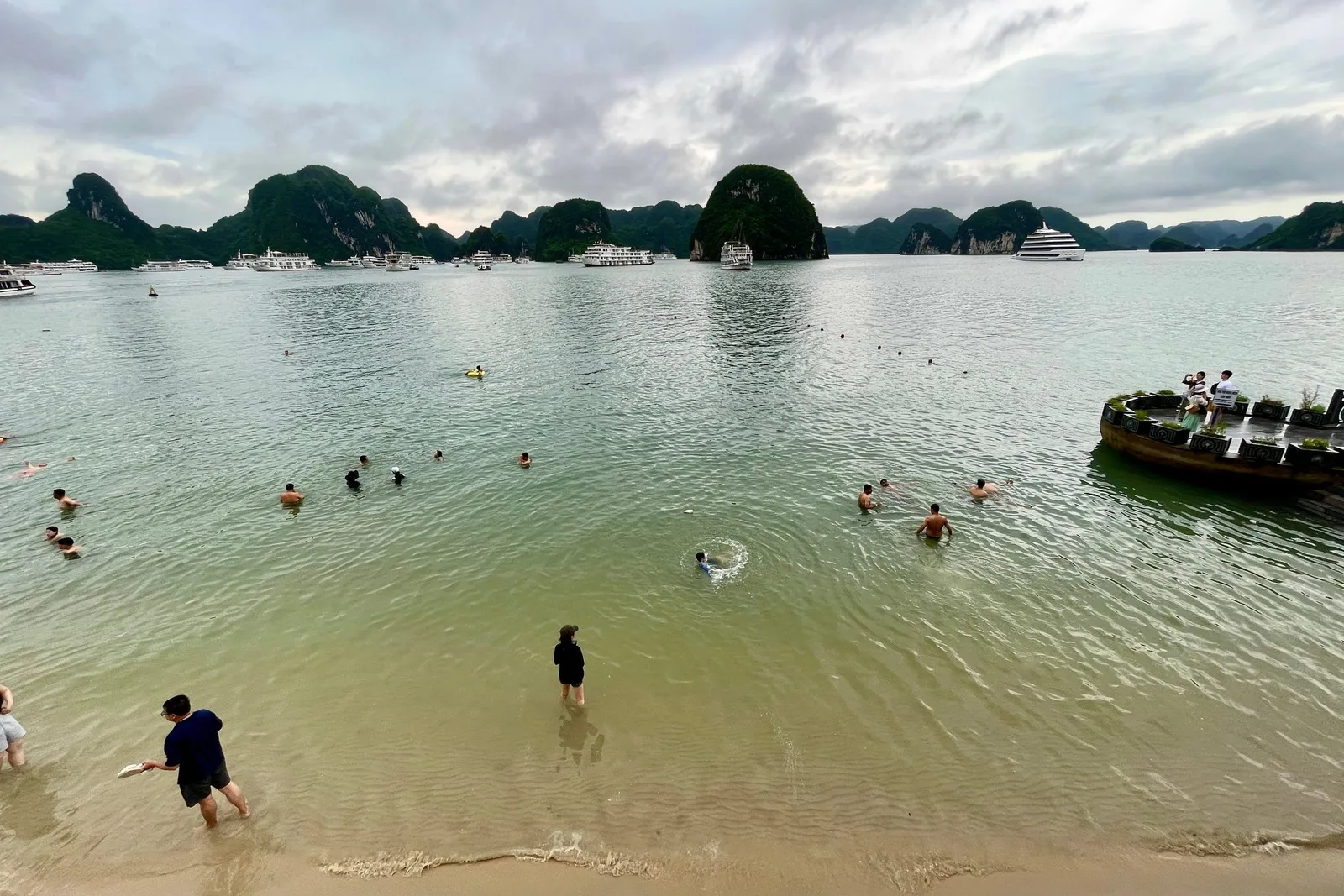 People swimming and wading in shallow water near a beach with limestone karsts and cruise ships in the background.