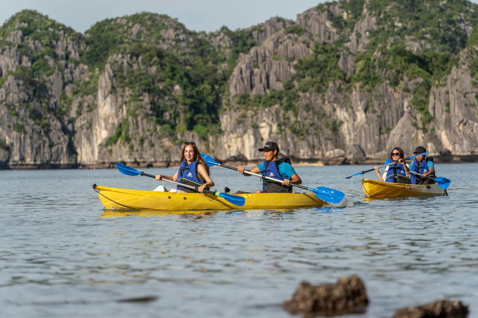 Four people kayaking in yellow kayaks with blue paddles on calm water in front of tall limestone karst formations.