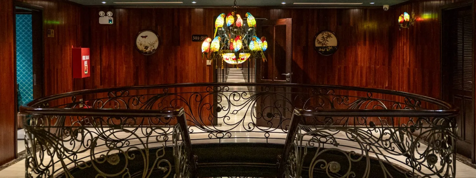 A wood-paneled stairwell landing with an ornate wrought-iron railing, a colorful parrot-shaped stained-glass chandelier, round decorative wall plaques, a red fire cabinet and a room-number sign reading 301-315.