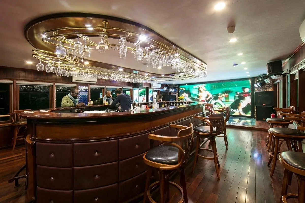 Interior of a wood-paneled bar lounge with an oval leather-fronted bar, hanging wine glasses, bar stools, a large LED screen, and three people at or behind the bar.