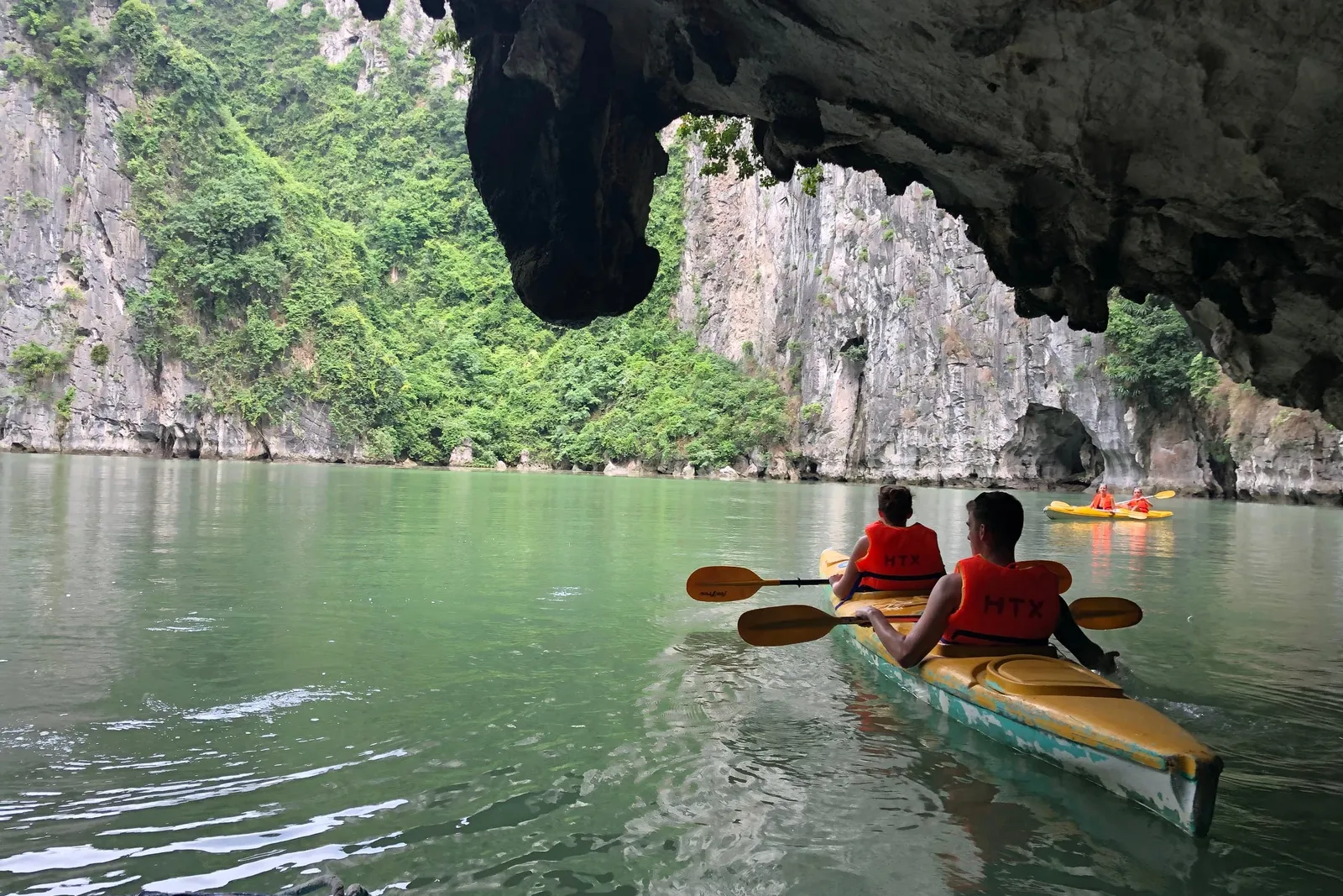 Two people kayaking in green water through a cave opening with limestone karsts and another kayak visible in the distance.